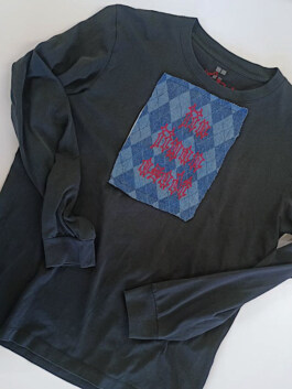 Schwarzes Langarmshirt mit rundem Ausschnitt und einem aufgenähten, blau-karierten Stoffquadrat mit roter, gotisch gestalteter Schrift auf der Brust. Black long-sleeve shirt with a round neckline and a sewn-on blue checkered fabric square featuring red Gothic-style text on the chest. 