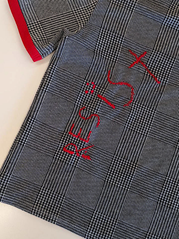 Schwarz-weiß kariertes T-Shirt mit rotem Saum und roter Stickerei 'RESIST' auf der Vorderseite. Black-and-white checkered T-shirt with red trim and red embroidered text 'RESIST' on the front.