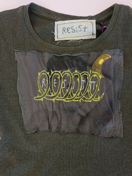 Dunkelgrünes T-Shirt mit kurzem Arm und rundem Ausschnitt, vorne ein schwarzes Stoffquadrat mit gelb-weißer Stickerei und einem gelben Halbmond. Innen am Kragen ein Etikett mit der Aufschrift 'RESIST'. Dark green short-sleeve T-shirt with round neckline, featuring a black fabric square on the front with yellow and white embroidery and a yellow crescent. Inside collar label reads 'RESIST'. 