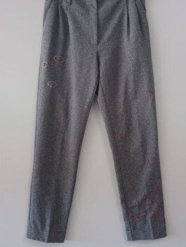Graue Hose mit mehreren roten Augenmotiven, die über die Beine verteilt sind, vor hellem Hintergrund. Gray pants with multiple red eye motifs scattered across the legs against a light background. 