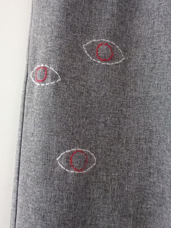 Grauer Stoff mit drei gestickten Augen in Weiß und Rot, die unregelmäßig angeordnet sind. Gray fabric with three embroidered eyes in white and red, arranged irregularly. 