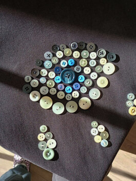 Verschiedene Knöpfe in mehreren Farben und Größen, angeordnet auf dunklem Stoff in Form eines Auges mit zwei Tropfen darunter. Various buttons in multiple colors and sizes arranged on dark fabric in the shape of an eye with two drops below. 