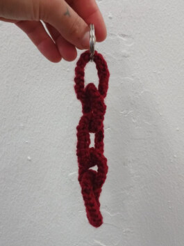 Hand hält einen Schlüsselanhänger aus dunkelrotem, gehäkeltem Garn mit drei Gliedern und einem Metallring. Hand holding a keychain made of dark red crocheted yarn with three links and a metal ring. 
