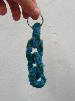 Hand hält einen Schlüsselring mit einer gehäkelten Kette aus blau-grünem Garn mit drei Gliedern. Hand holding a key ring with a crocheted chain of three links in blue and green yarn. 