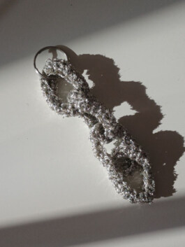Silberfarbener Schlüsselanhänger aus grob gehäkeltem Garn mit Metallring, liegt auf hellgrauer Oberfläche mit Schatten. Silver-colored keychain made of loosely crocheted yarn with metal ring, lying on light gray surface with shadow. 