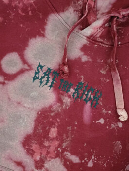 Nahaufnahme eines roten Kapuzenpullovers mit hellen Flecken und einem grünen, stilisierten Schriftzug in der Mitte Close-up of a red hoodie with light spots and a green stylized text in the center