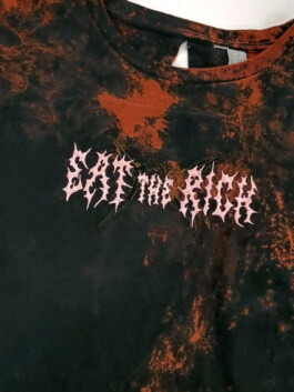 Nahaufnahme eines schwarz-orange gefleckten T-Shirts mit dem Schriftzug 'EAT THE RICH' in verzerrter Schrift auf der Brust Close-up of a black and orange speckled T-shirt with the distorted text 'EAT THE RICH' on the chest 