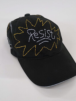 Schwarze Baseballkappe mit der weißen Stickerei 'Resist' in einer gelben, gezackten Umrandung auf der Vorderseite Black baseball cap with white embroidered word 'Resist' inside a yellow jagged outline on the front 