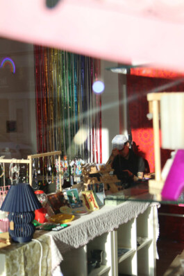 Sales table with jewelry, small artworks, and a blue lamp on a white-covered table in front of a background with colorful vertical strips. Verkaufsstand mit Schmuck, kleinen Kunstwerken und einer blauen Lampe auf einem mit weißer Decke bedeckten Tisch vor einem Hintergrund mit bunten, vertikalen Lamellen. 
