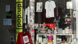 Shop window displaying T-shirts, posters, stickers, and various art objects including a red sandwich board sign reading 'BROKE ARTIST SHOP' Schaufenster eines Ladens mit T-Shirts, Postern, Aufklebern und verschiedenen Kunstobjekten, darunter ein rotes Aufsteller-Schild mit der Aufschrift 'BROKE ARTIST SHOP' 