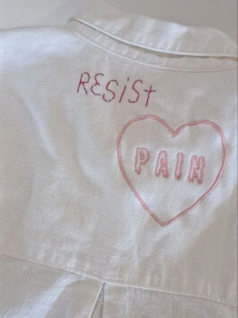 Rückseite eines weißen ärmellosen Jeanshemds mit den aufgestickten Worten 'RESIST' und 'PAIN' in einem Herz