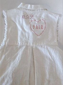 Rückseite eines weißen ärmellosen Jeanshemds mit den aufgestickten Worten 'RESIST' und 'PAIN' in einem Herz