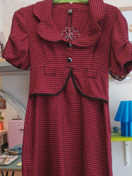 Rotes Kleid mit schwarzem Karomuster, kurzen Puffärmeln, abgerundetem Kragen, zwei schwarzen Knöpfen vorne und schwarzem Besatz am Saum des Oberteils 