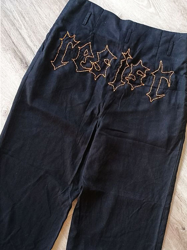 Schwarze Hose mit oranger Stickerei in gotischer Schrift, die das Wort 'resist' bildet